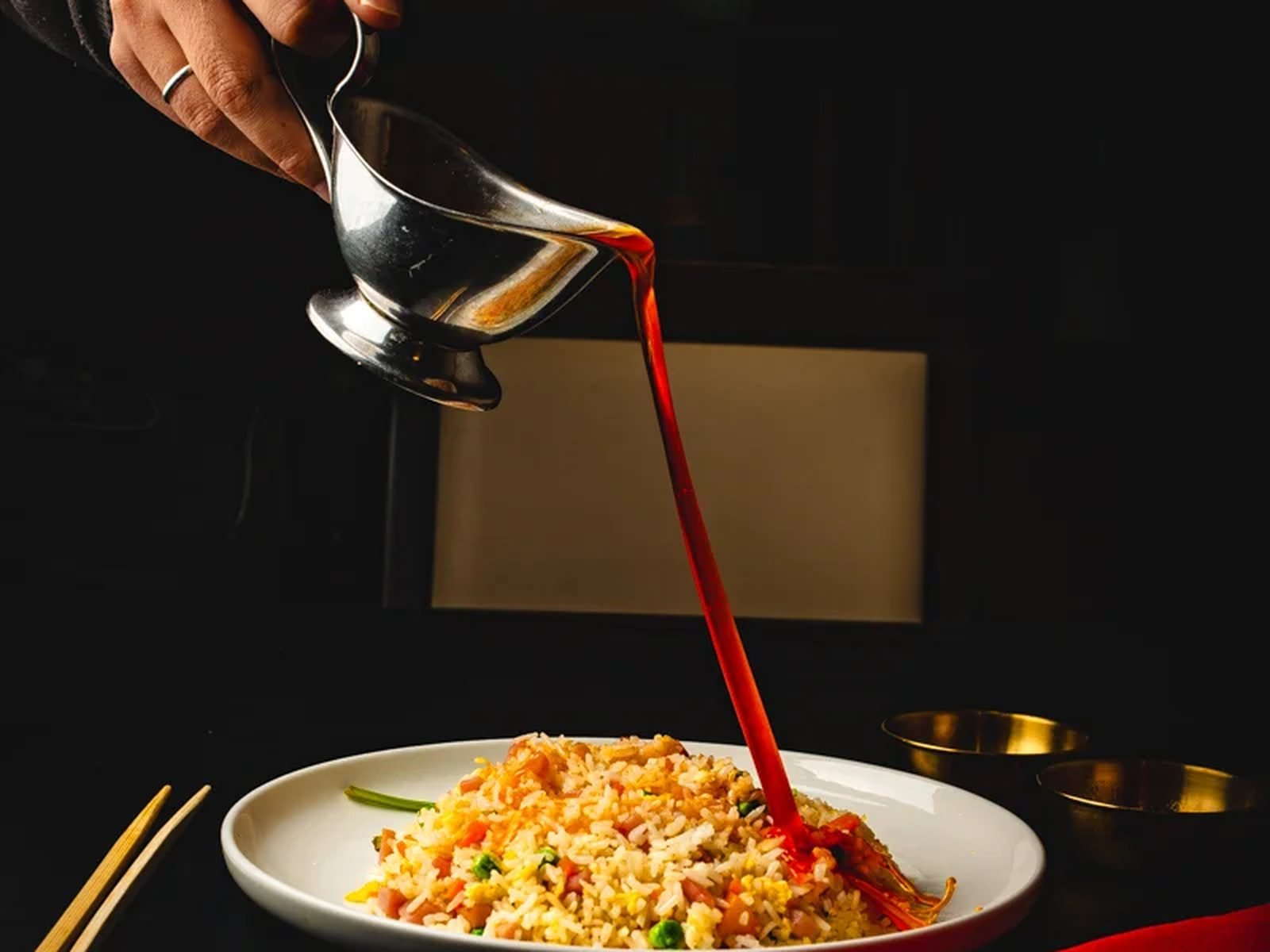 Mano vertiendo salsa roja sobre arroz frito en plato blanco con fondo negro