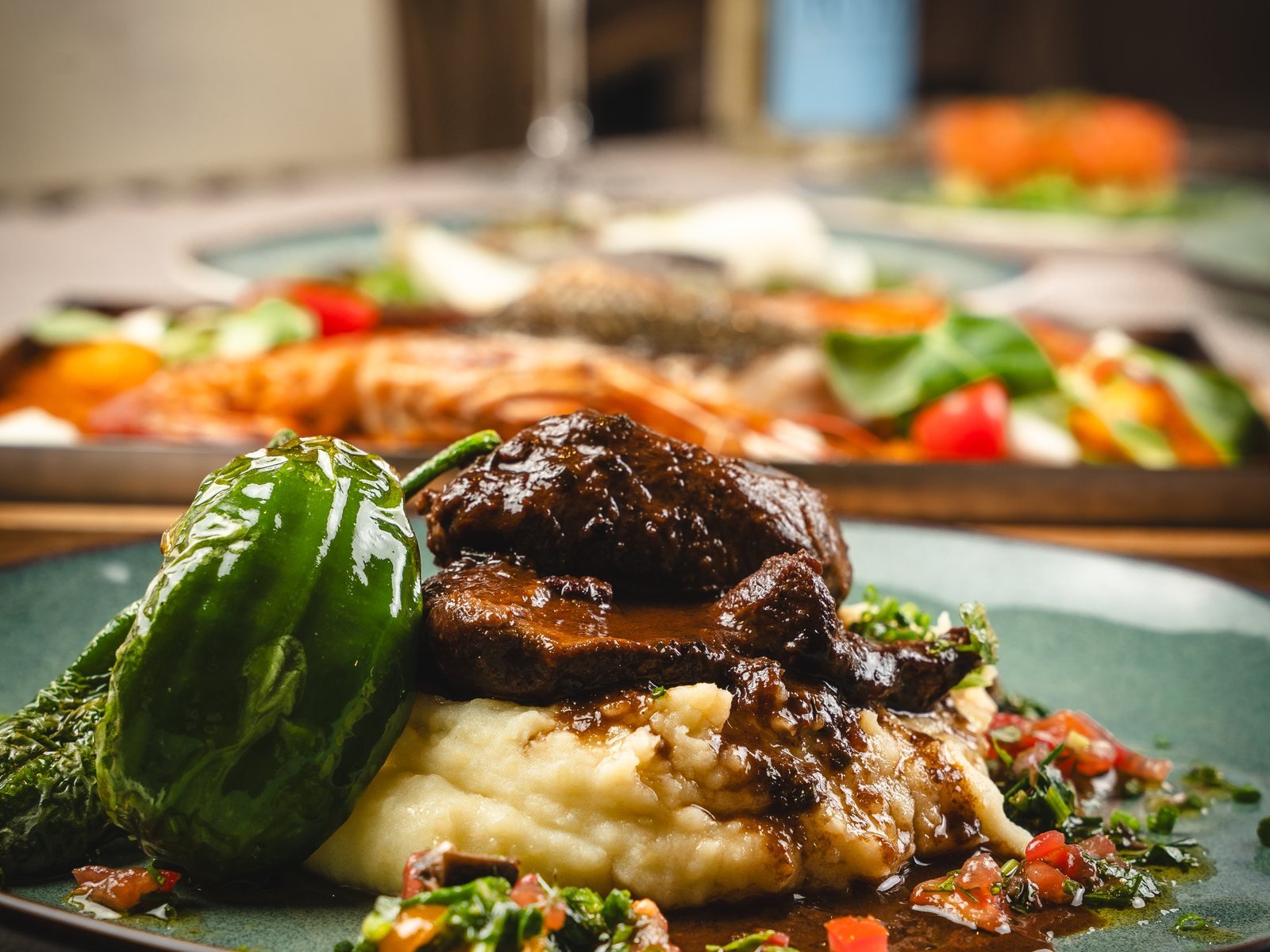 Carrillera con puré y pimiento verde en plato verde, con más platos en la mesa de fondo, fotos que explican cuánto cuesta un fotógrafo gastronómico según la carta