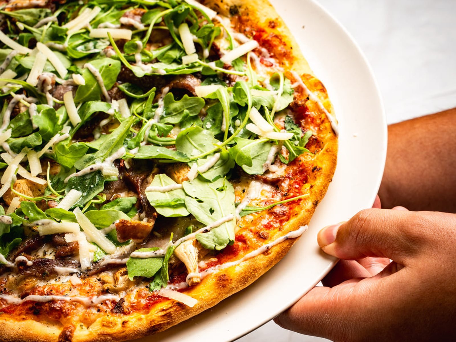 Mano sirviendo una pizza con rúcula y parmesano en plato blanco