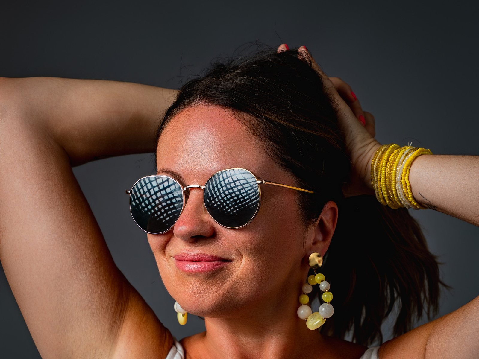 Retrato de una mujer sonriente con gafas de sol redondas, pendientes y pulseras amarillas en estudio